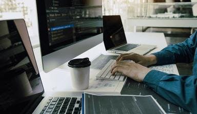 Person working on a desk with a couple of laptops, computer and a cup of coffee.